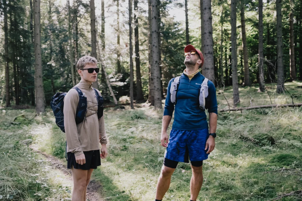 Zwei Menschen im Wald während einer Wanderung. Der Mann links trägt Sonnenbrille und einen Rucksack, während der Mann rechts eine Kappe trägt und nach oben schaut. Um sie herum sind hohe Bäume und grünes Unterholz.