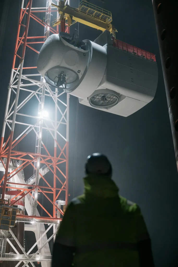 Ein Arbeiter in Schutzkleidung beobachtet eine Gondel, die von einem Kran während der Nacht gehoben wird. Die Szene ist gut beleuchtet und zeigt die Technik und Infrastruktur des Aufbaus.